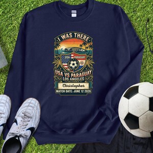 Navy blue sweatshirt mockup featuring a personalized vintage-style “I Was There” USA vs Paraguay soccer match design. Graphic includes a sunset stadium scene, palm trees, soccer ball, USA and Paraguay flags, “Los Angeles California,” custom name “Christopher,” and “Match Date: June 12 2026.” Displayed flat lay on green grass with soccer cleats and a soccer ball, perfect personalized souvenir sweatshirt for soccer fans, match day, and unforgettable sports memories.