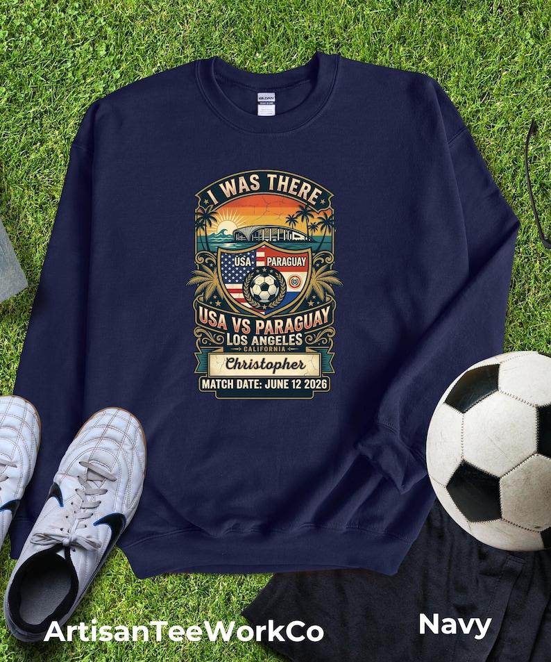 Navy blue sweatshirt mockup featuring a personalized vintage-style “I Was There” USA vs Paraguay soccer match design. Graphic includes a sunset stadium scene, palm trees, soccer ball, USA and Paraguay flags, “Los Angeles California,” custom name “Christopher,” and “Match Date: June 12 2026.” Displayed flat lay on green grass with soccer cleats and a soccer ball, perfect personalized souvenir sweatshirt for soccer fans, match day, and unforgettable sports memories.