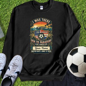 Black sweatshirt mockup featuring a personalized vintage-style “I Was There” USA vs Paraguay soccer match design. Graphic includes a sunset stadium scene, palm trees, soccer ball, USA and Paraguay flags, “Los Angeles California,” customizable name text, and “Match Date: June 12 2026.” Displayed flat lay on green grass with soccer cleats and a soccer ball, perfect personalized souvenir sweatshirt for soccer fans, match day, travel memories, and sports lovers.