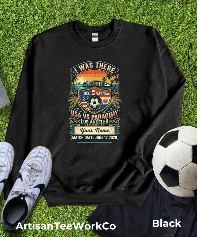 Black sweatshirt mockup featuring a personalized vintage-style “I Was There” USA vs Paraguay soccer match design. Graphic includes a sunset stadium scene, palm trees, soccer ball, USA and Paraguay flags, “Los Angeles California,” customizable name text, and “Match Date: June 12 2026.” Displayed flat lay on green grass with soccer cleats and a soccer ball, perfect personalized souvenir sweatshirt for soccer fans, match day, travel memories, and sports lovers.