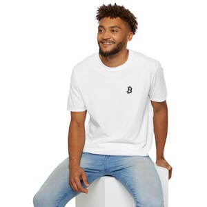 May include: White t-shirt with a small black Bitcoin symbol on the chest. The shirt has short sleeves and a crew neck. The person in the image is wearing blue jeans.