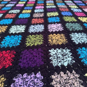Peut inclure: Une couverture au crochet multicolore faite à la main. La couverture présente un fond noir avec de nombreux carrés de différentes couleurs, dont le rouge, le bleu, le violet et le jaune. Les carrés ont un motif texturé en forme de fleur.