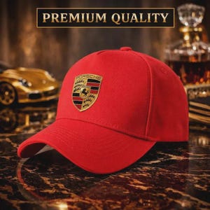 May include: A red baseball cap with the Porsche logo on the front. The cap has a curved brim and the text "PREMIUM QUALITY" above it. The Porsche logo is a gold, black, and red emblem.