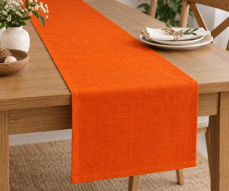 K&ouml;nnte beinhalten: Ein leuchtend orangefarbener Tischl&auml;ufer mit sichtbarer Webstruktur, der der L&auml;nge nach &uuml;ber einen Holztisch drapiert ist. Der Tischl&auml;ufer hat saubere, gerade Kanten und einen einfachen ges&auml;umten Abschluss, der eine einheitliche Farbfl&auml;che bildet.