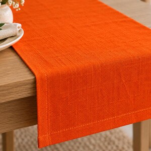 K&ouml;nnte beinhalten: Ein rechteckiger orangefarbener Tischl&auml;ufer mit sichtbarer Webstruktur und gen&auml;hten ges&auml;umten Kanten. Der Stoff ist einfarbig orange und liegt flach auf der Oberfl&auml;che eines Holztisches, wobei er &uuml;ber die Kante mit einem sauberen Abschluss ragt.