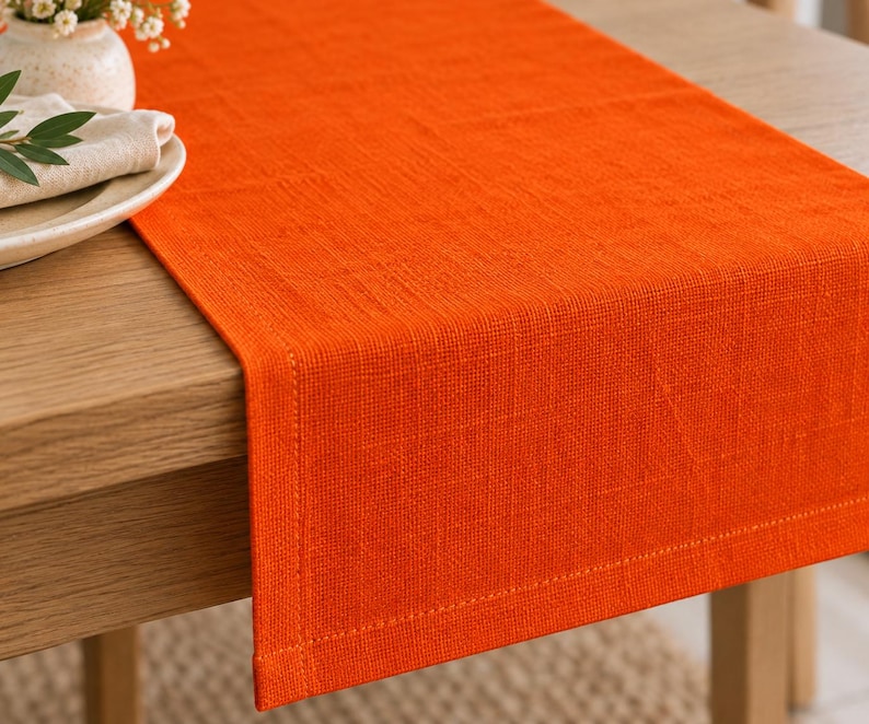 K&ouml;nnte beinhalten: Ein rechteckiger orangefarbener Tischl&auml;ufer mit sichtbarer Webstruktur und gen&auml;hten ges&auml;umten Kanten. Der Stoff ist einfarbig orange und liegt flach auf der Oberfl&auml;che eines Holztisches, wobei er &uuml;ber die Kante mit einem sauberen Abschluss ragt.