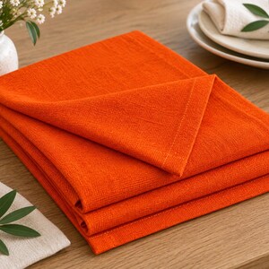 K&ouml;nnte beinhalten: Ein Stapel aus drei gefalteten, quadratischen Servietten in leuchtendem Orange. Die Servietten weisen eine sichtbare Webstruktur und saubere, vern&auml;hte Kanten auf. Der Stoff wirkt wie ein mittelschweres Textil mit matter Oberfl&auml;che, ordentlich &uuml;bereinandergelegt.