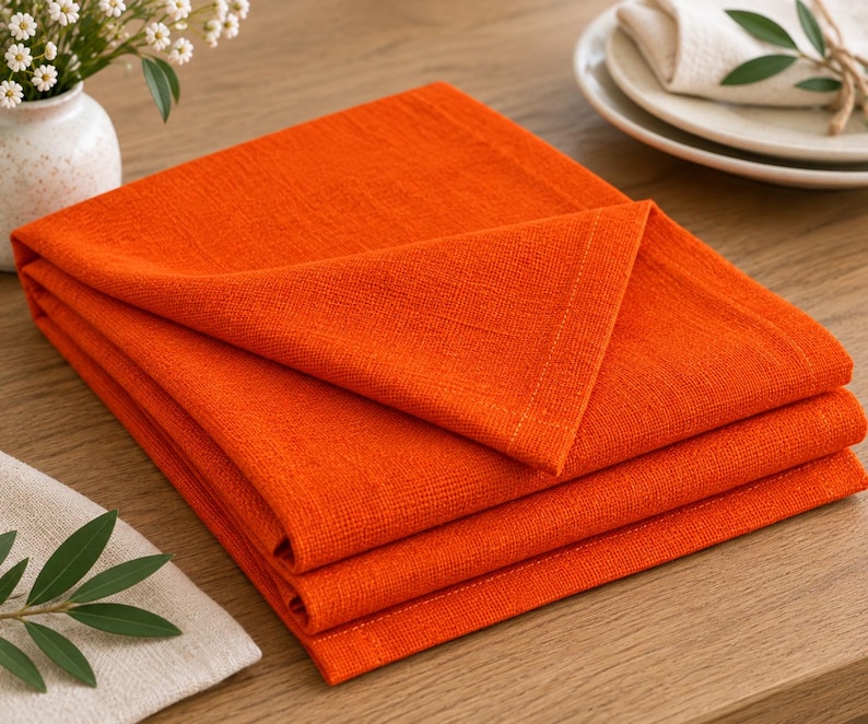 K&ouml;nnte beinhalten: Ein Stapel aus drei gefalteten, quadratischen Servietten in leuchtendem Orange. Die Servietten weisen eine sichtbare Webstruktur und saubere, vern&auml;hte Kanten auf. Der Stoff wirkt wie ein mittelschweres Textil mit matter Oberfl&auml;che, ordentlich &uuml;bereinandergelegt.