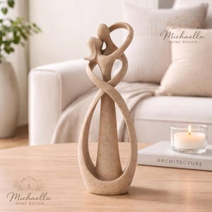 Könnte beinhalten: Eine beige abstrakte Skulptur, die zwei sich umarmende Figuren mit verschlungenen Armen und Körpern darstellt. Die Skulptur steht auf einem Holztisch, mit einer brennenden Kerze und einem Buch mit dem Titel "ARCHITECTURE" im Hintergrund. Ein Dekorationsartikel.