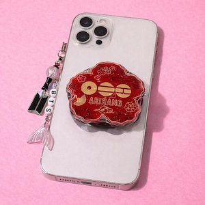 Puede incluir: Un smartphone blanco con un agarre para teléfono rojo en forma de flor con el texto "ARIRANG". Un dije con las letras "BTS" y una cola de sirena cuelga del teléfono. El teléfono está sobre una superficie rosa.