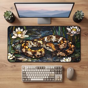 May include: A rectangular desk mat featuring a stained glass style illustration of a brown and tan patterned snake coiled among white water lilies and green lily pads on a dark pond surface, positioned on a wooden desk with a keyboard and mouse.