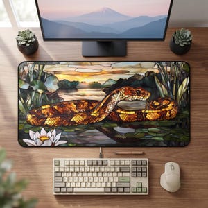 Könnte beinhalten: Eine rechteckige Schreibtischunterlage mit einer Illustration im Buntglas-Stil, die eine braun-orange Schlange zeigt, die sich zwischen grünen Seerosenblättern und einer weißen Seerose windet. Der Hintergrund zeigt einen Sonnenuntergang über Wasser.