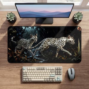 Peut inclure: Un tapis de bureau rectangulaire présentant une illustration détaillée de deux chats sauvages tachetés marchant dans une forêt sombre et dense avec des ombres profondes et du feuillage. Le tapis est posé sur une surface en bois sous un écran.