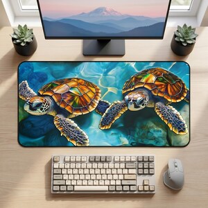 May include: A rectangular desk mat featuring a vibrant illustration of two sea turtles swimming in clear blue water. The turtles have detailed, multi-coloured shells in shades of orange, yellow, and green, with dark patterned flippers.