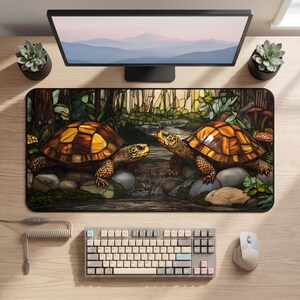 Op de afbeelding: Een rechthoekige bureaumat met een illustratie in glas-in-loodstijl van twee schildpadden die tegenover elkaar staan bij een beekje. De schildpadden hebben barnsteenkleurige schilden en een getextureerde huid, omringd door rotsen, groen gebladerte en kleine rode paddenstoelen.