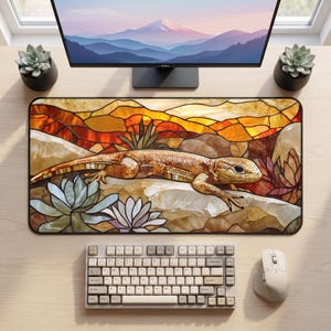 Pode incluir: Um tapete de secretária retangular com uma ilustração em estilo de vitral de um lagarto a descansar sobre rochas entre suculentas. O design incorpora tons quentes de laranja, amarelo e castanho com uma paisagem de montanha e uma borda preta.