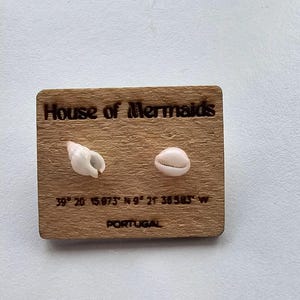 Può includere: Targa in legno con la scritta "House of Mermaids" e due conchiglie. La targa include anche le coordinate 39° 20 15.873" N 9° 21 36.583" W e la parola "PORTUGAL".