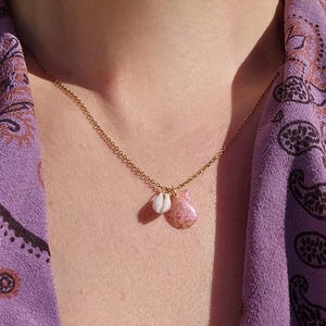 May include: A gold-coloured necklace with two pendants: a small, white seashell and a pink, teardrop-shaped stone. The necklace is set against a purple paisley patterned garment.