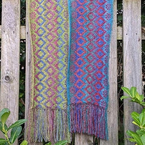 May include: Two woven scarves with diamond patterns and fringe hang on a wooden fence. One scarf features green, purple, and yellow hues, while the other displays blue, red, and purple colors. The scarves are likely handmade.