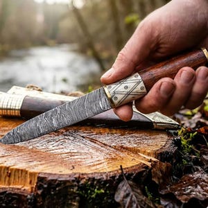 Op de afbeelding: Een handgemaakt mes met een patroon op het lemmet en een houten handvat met bot- en messingaccenten. Het mes ligt op een boomstronk, met de schede op de achtergrond. Het lemmet heeft een uniek, wervelend patroon.