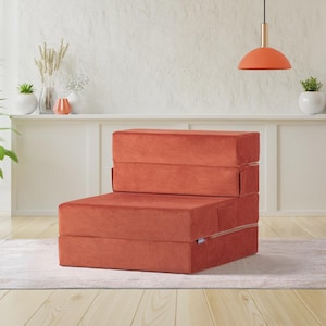 Könnte beinhalten: Ein ziegelroter, faltbarer Bodenstuhl. Der Stuhl besteht aus einem weichen, samtigen Material und ist zu einer kompakten, stuhlähnlichen Form gefaltet. Der Stuhl steht auf einem hellen Teppich in einem Raum mit weißer Wand.