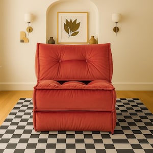 Könnte beinhalten: Ein moderner, rot-orangefarbener Polstersessel mit gesteppter Rückenlehne und Sitzfläche. Der Sessel steht auf einem schwarz-weiß karierten Teppich. Der Hintergrund umfasst einen gerahmten botanischen Druck und Wandleuchten.