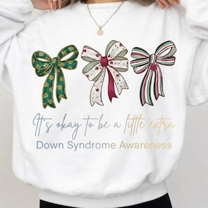 May include: White sweatshirt with three decorative bows in green, red, and white. The bows have different patterns. The text "It's okay to be a little extra" and "Down Syndrome Awareness" are printed below the bows.