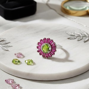 May include: A silver ring with a flower-shaped design. The ring features a large, oval, light green center stone surrounded by smaller, rectangular, pink stones. Several loose gemstones in shades of pink and green are scattered around the ring.
