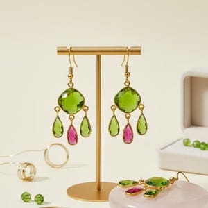 Puede incluir: Pendientes dorados con grandes piedras preciosas verdes y rosas expuestos en un soporte dorado. Un par de pendientes están en una caja de joyería blanca. Joyas y cuentas adicionales están dispersas.