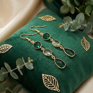 May include: Gold-toned earrings with green gemstones and a teardrop shape, displayed on a green velvet cushion. Decorative gold leaf charms are scattered around the earrings, with eucalyptus leaves in the background.
