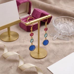 Può includere: Espositore per orecchini dorato con due paia di orecchini pendenti. Ogni orecchino presenta una montatura dorata con gemme rosa, azzurre e ovali blu grandi. Sono visibili anche un portagioie in velluto e una scheda vuota.