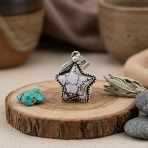 May include: A star-shaped pendant crafted from sterling silver featuring a white stone with dark brown veining. The pendant has a beaded edge and a small silver leaf detail at the top, attached to a polished silver bail for hanging on a chain.