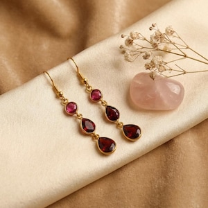 May include: A pair of gold-coloured drop earrings, each with three red gemstones. The top stone is round, and the lower two are teardrop-shaped. A pink stone and small white flowers are in the background.