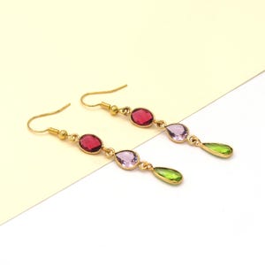 Peut inclure: Une paire de boucles d'oreilles pendantes dorées. Chaque boucle d'oreille comporte trois pierres précieuses colorées : un ovale rouge, un ovale lavande et une goutte verte. Les boucles d'oreilles sont présentées sur un fond blanc et jaune.