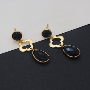 Op de afbeelding: Een paar goudkleurige oorhangers met zwarte edelstenen. Elke oorbel heeft een ronde stud, een klavervormig accent en een druppelvormige steen. De oorbellen worden gepresenteerd tegen een zwart-witte achtergrond.