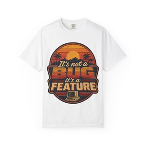 May include: A white short-sleeve t-shirt featuring a circular graphic with a retro sunset, palm trees, and mountains. The text reads It's not a BUG it's a FEATURE above a small illustration of a vintage computer terminal in orange and blue.