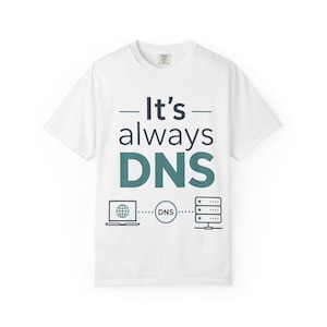 Può includere: Una t-shirt bianca a maniche corte con la scritta It's always DNS in blu scuro e verde acqua. Sotto il testo, un grafico mostra un computer portatile collegato a un cerchio DNS, a sua volta collegato a un'icona di server, illustrando un flusso di rete.