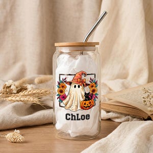 May include: A clear glass tumbler with a bamboo lid and a metal straw. The glass features a printed graphic of a ghost wearing an orange witch hat, a black cat, a pumpkin, and colourful flowers. The name Chloe is printed in black below the graphic.