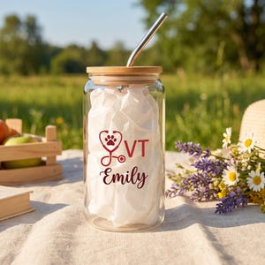 Puede incluir: Un vaso transparente con tapa de bambú y pajita de metal. El vaso presenta un gráfico rojo de un estetoscopio con una huella de pata en el centro, las letras VT y el nombre Emily impreso en letra cursiva. El vaso está relleno de papel blanco.