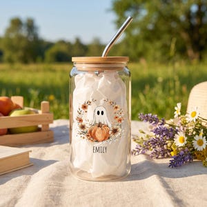 May include: A clear glass tumbler with a bamboo lid and a metal straw. The glass features a printed design of a white ghost sitting on an orange pumpkin, surrounded by a wreath of autumn flowers and the name EMILY printed in black text.