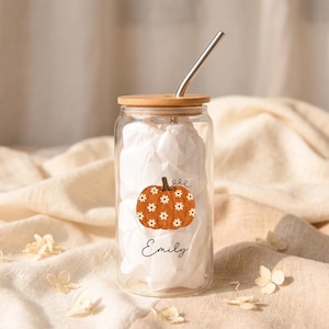 May include: A clear glass tumbler with a bamboo lid and a metal straw. The glass features a graphic of an orange pumpkin decorated with small white flowers and the name Emily written in black cursive script below the pumpkin illustration.