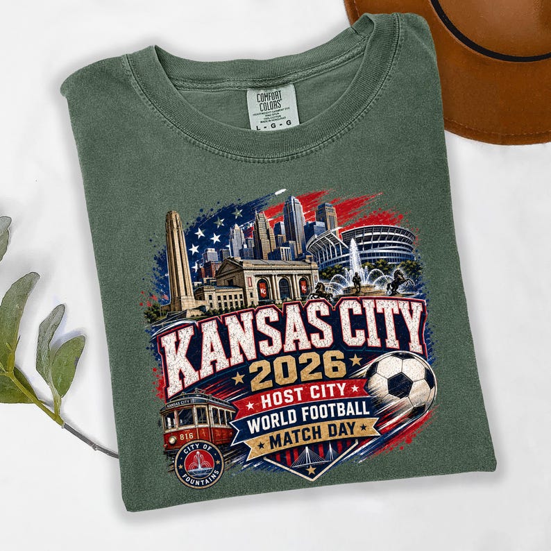 May include: A sage green t-shirt featuring a graphic of the Kansas City skyline, a red streetcar, and a soccer ball. The text reads Kansas City 2026 Host City World Football Match Day, set against a stylized red, white, and blue background design.