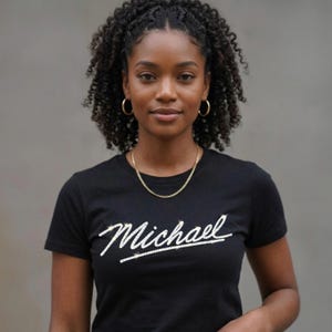 May include: Black crew neck t-shirt with the name "Michael" in a white, cursive font. The shirt is worn with a gold chain necklace and gold hoop earrings. The person has curly dark hair.