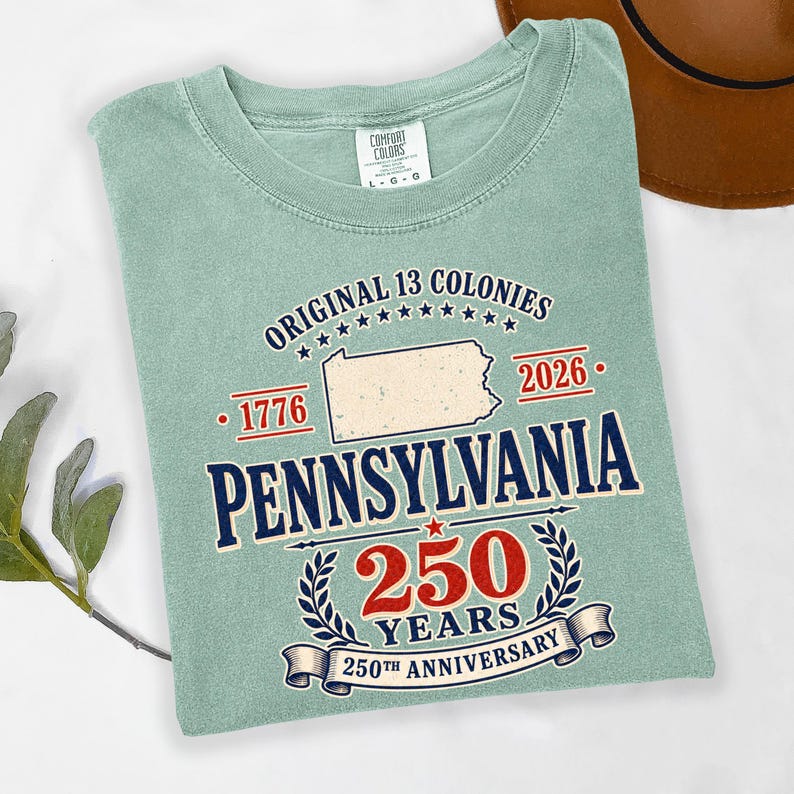 May include: A light green t-shirt with the text "ORIGINAL 13 COLONIES" and "PENNSYLVANIA" in navy blue. The shirt also features the numbers "1776" and "2026", and "250 YEARS 250TH ANNIVERSARY" in red and navy blue.