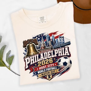 May include: A cream-colored t-shirt featuring a graphic of the Liberty Bell, the Philadelphia skyline, and a soccer ball. The text reads PHILADELPHIA 2026 HOST CITY WORLD FOOTBALL MATCH DAY. The design incorporates red, white, and blue colors.