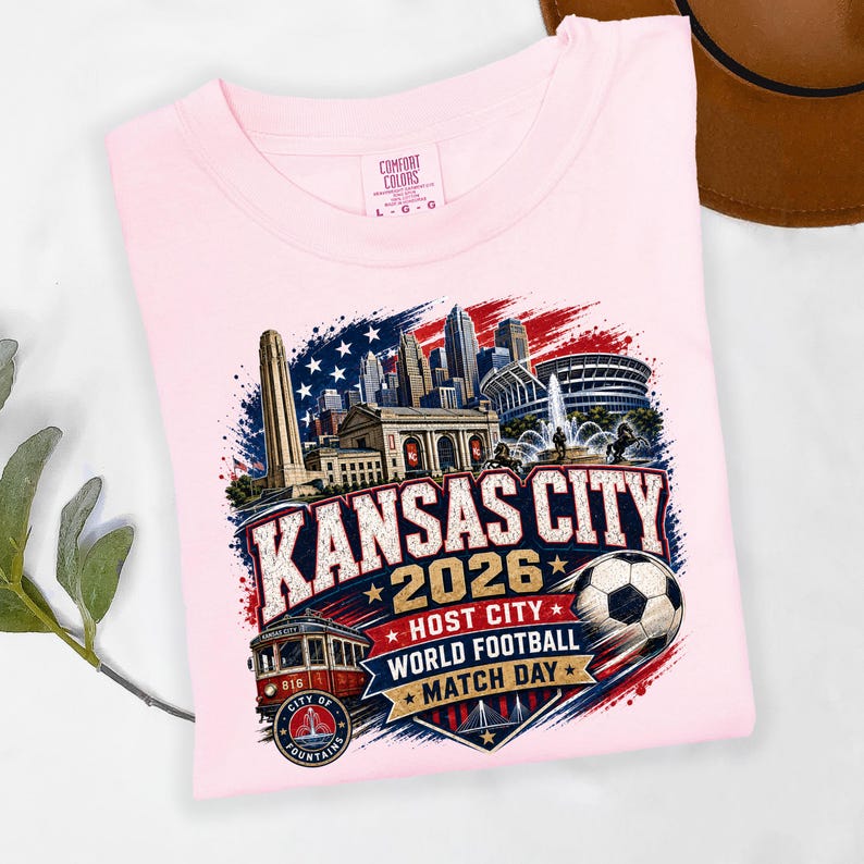 May include: A light pink t-shirt featuring a graphic of the Kansas City skyline, a streetcar, and a soccer ball. The text reads KANSAS CITY 2026 HOST CITY WORLD FOOTBALL MATCH DAY. The design includes red, white, and blue accents with stars.