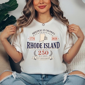 May include: A cream-colored t-shirt featuring the text "ORIGINAL 13 COLONIES" and "RHODE ISLAND" in navy blue. The shirt commemorates the 250th anniversary of Rhode Island, with the years 1776 and 2026.