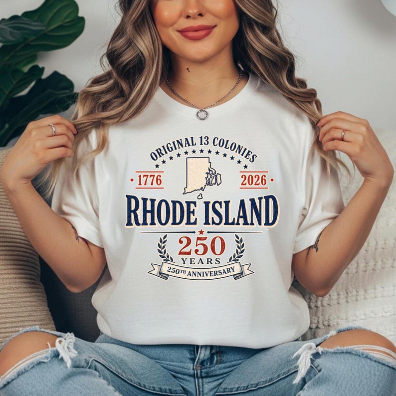May include: A cream-colored t-shirt featuring the text "ORIGINAL 13 COLONIES" and "RHODE ISLAND" in navy blue. The shirt commemorates the 250th anniversary of Rhode Island, with the years 1776 and 2026.