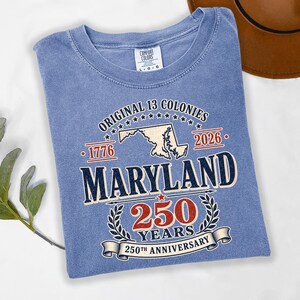May include: A light blue t-shirt with the text "ORIGINAL 13 COLONIES" and "MARYLAND 250 YEARS 250TH ANNIVERSARY". The shirt has a crew neck and short sleeves. The shirt is a size L-G-G.