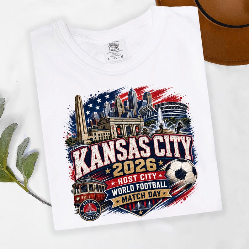 May include: A white t-shirt featuring a graphic print of the Kansas City skyline, a red streetcar, and a soccer ball. The text reads KANSAS CITY 2026 HOST CITY WORLD FOOTBALL MATCH DAY and CITY OF FOUNTAINS, set against a red, white, and blue background.